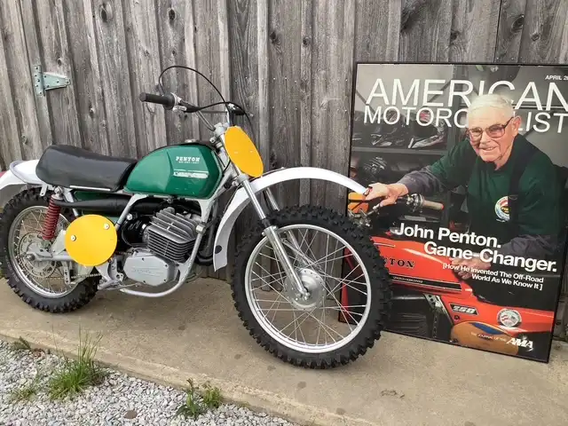 Vintage motorcycle beside John Penton poster outdoors.