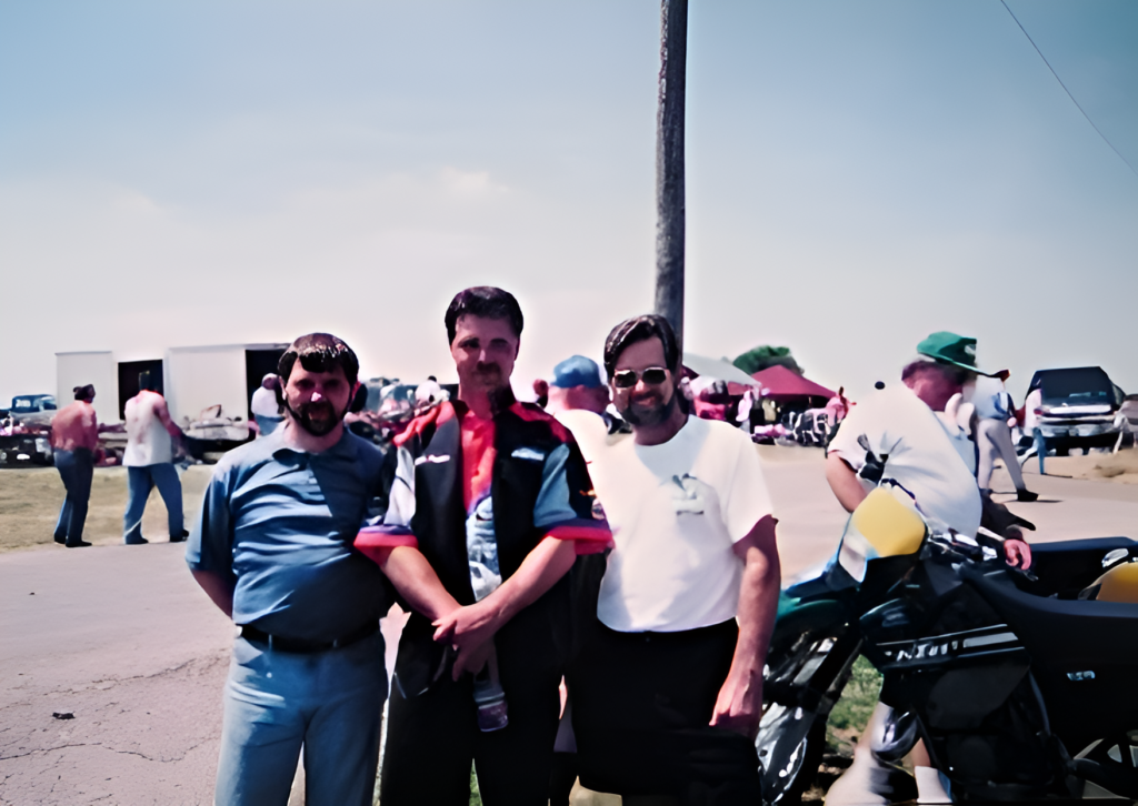Three men posing at outdoor event with motorcycles.
