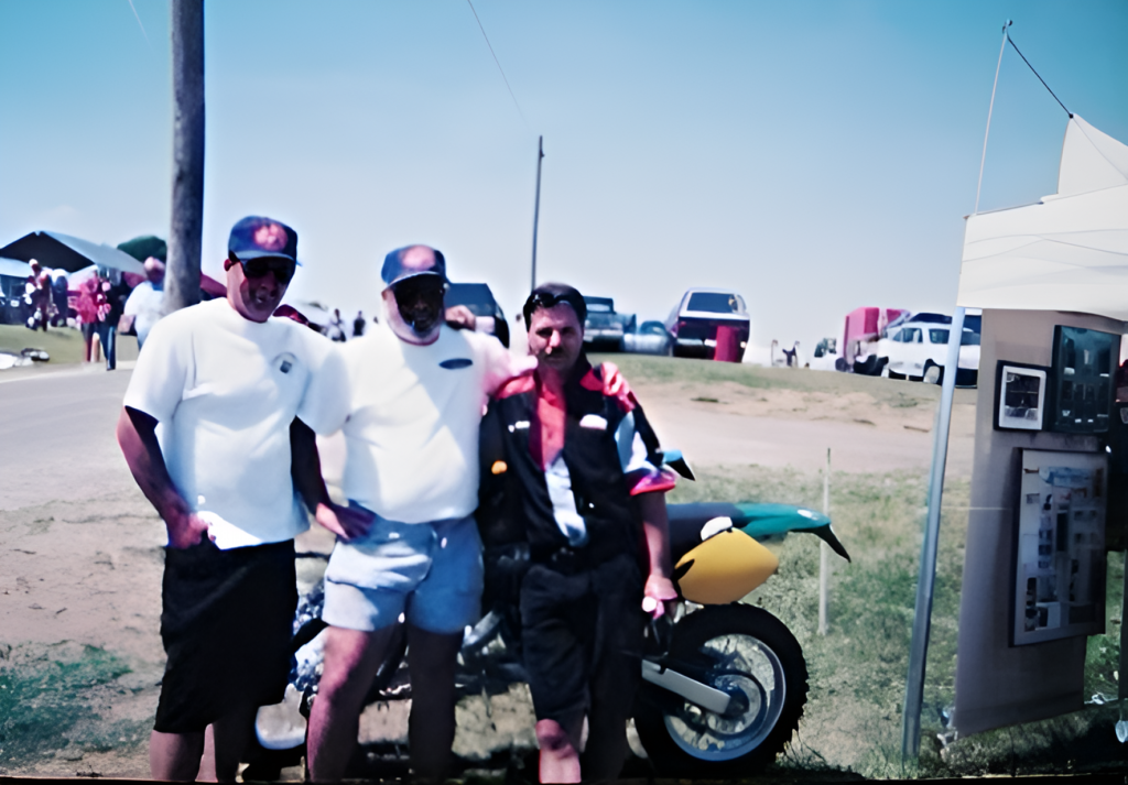 Three men posing with a motorcycle at outdoor event.