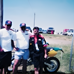 Three men posing with a motorcycle at outdoor event.