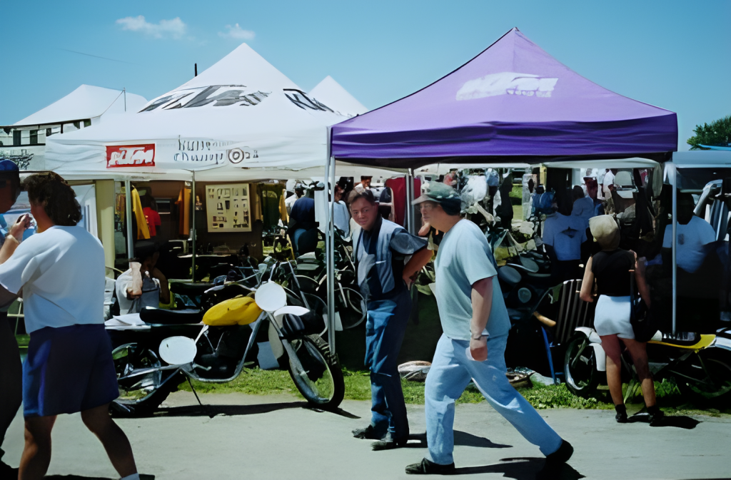 Outdoor market with tents and people browsing motorcycles.