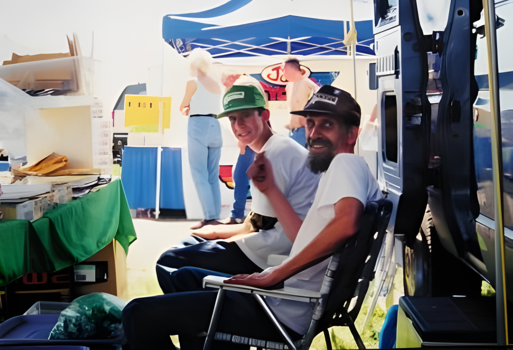 Two men sitting at outdoor market stand.