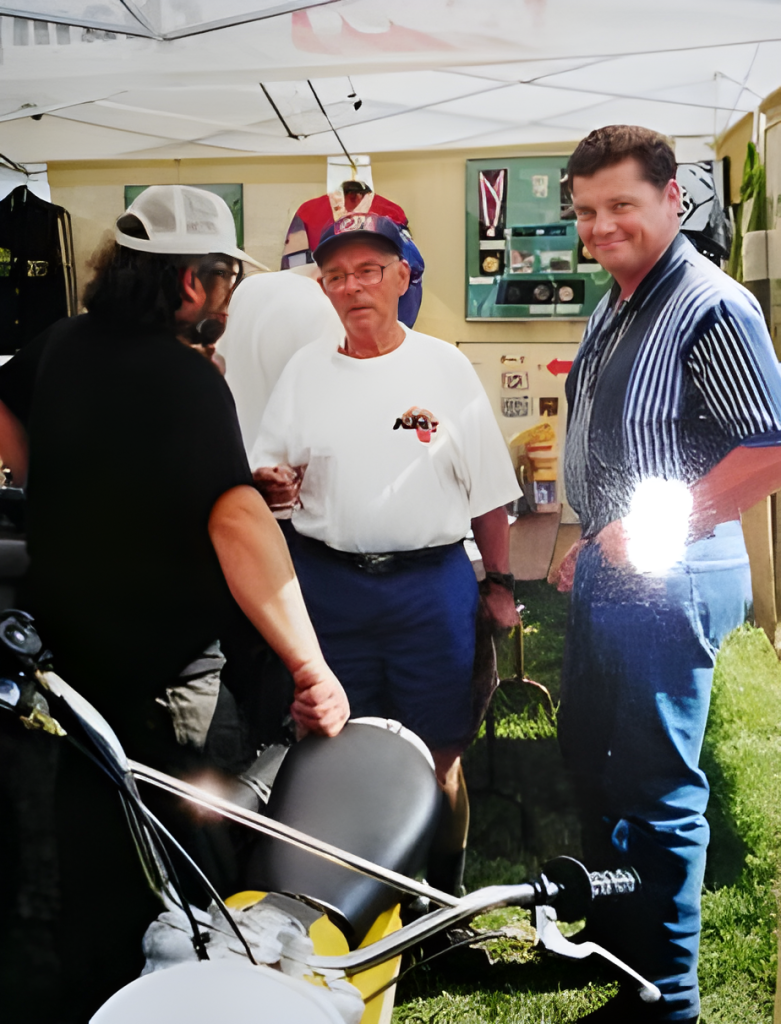 Three men talking at outdoor event near motorcycle.