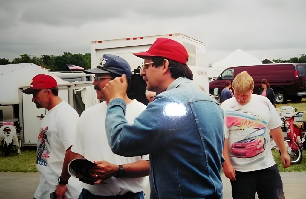Men at outdoor event with cars in background.