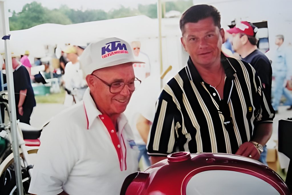 Two men smiling at outdoor event, motorcycle visible.