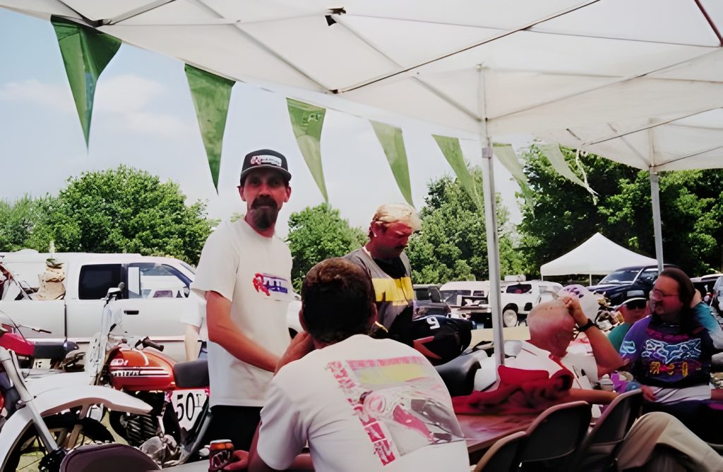 Motorcyclists gather under canopy at outdoor event.