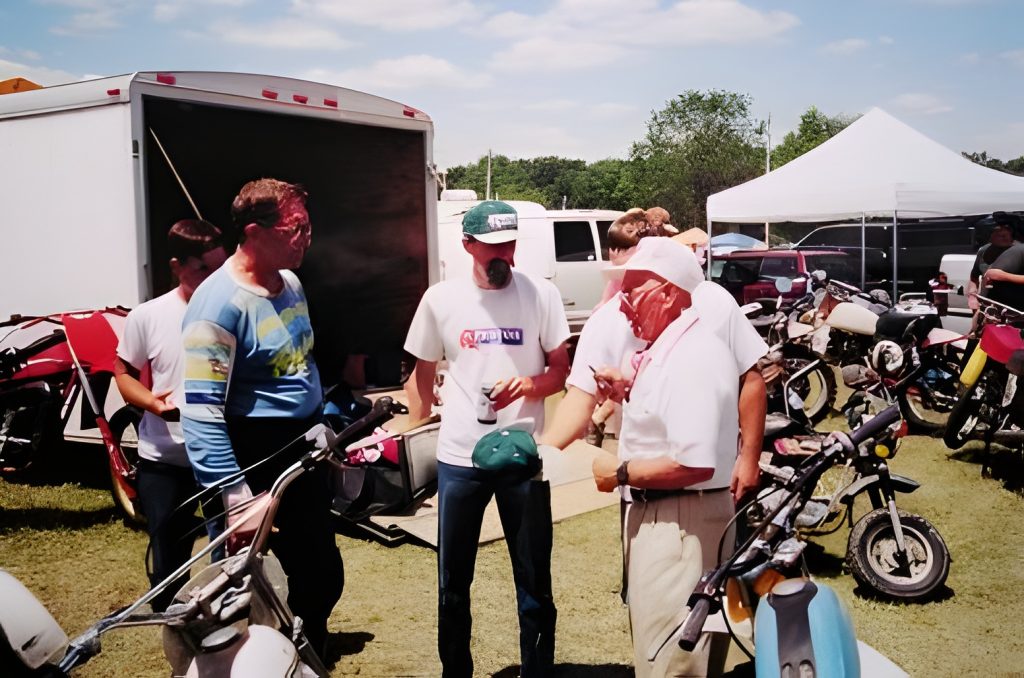 People gather around vintage motorcycles outside trailer.