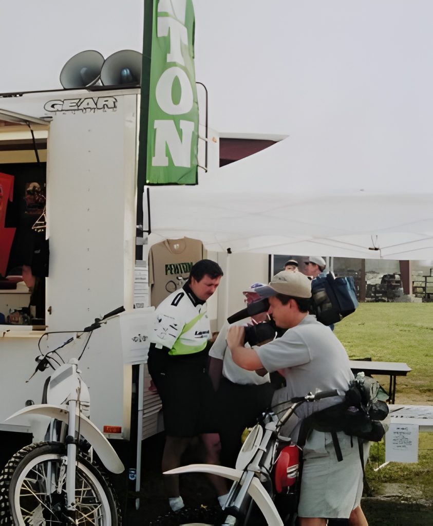Motorcyclist at outdoor event booth with camera bag.