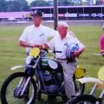 Two men with motorcycles at a racetrack event.