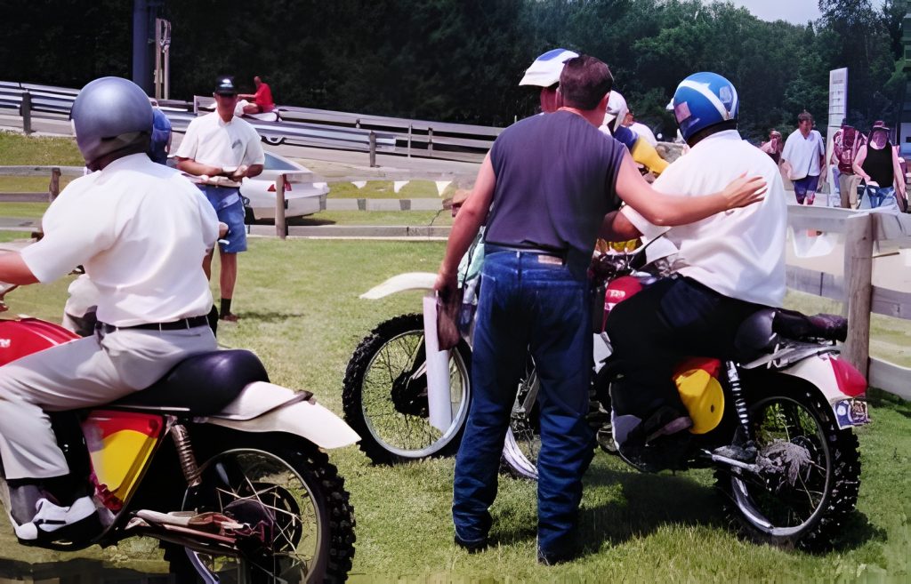 Group gathered around dirt bikes on grass field.
