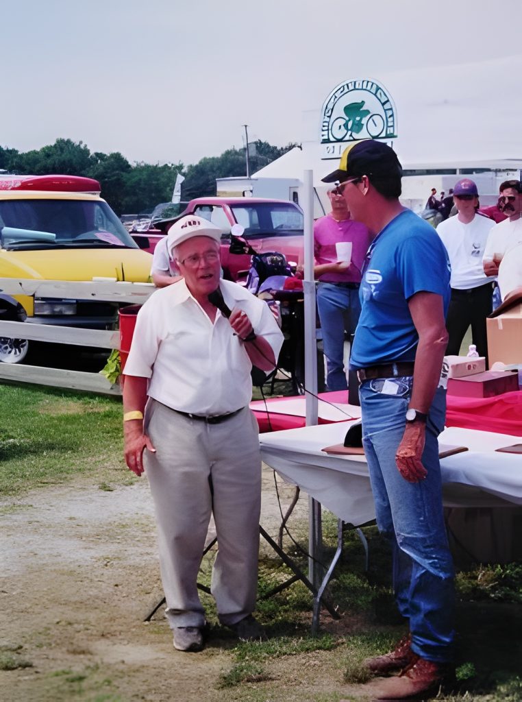 Two men talking at outdoor event near vehicles.