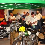 Group discussing vintage motorcycles under green tent.