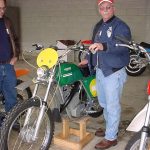 Two men examine vintage motorcycles in a garage.