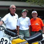 Three people smiling beside motorcycles outdoors.