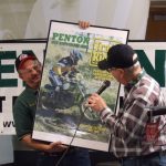 Two men holding Penton motorcycle poster at event.