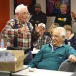 Elderly man speaking to seated group in classroom.
